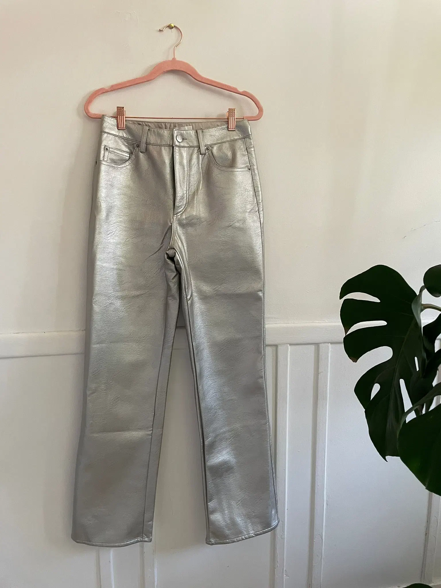 Pantalon-h&m-argent-visuel3 Pantalon argenté vegan de H&M suspendu sur un cintre rose, présenté contre un mur blanc minimaliste avec une plante en arrière-plan